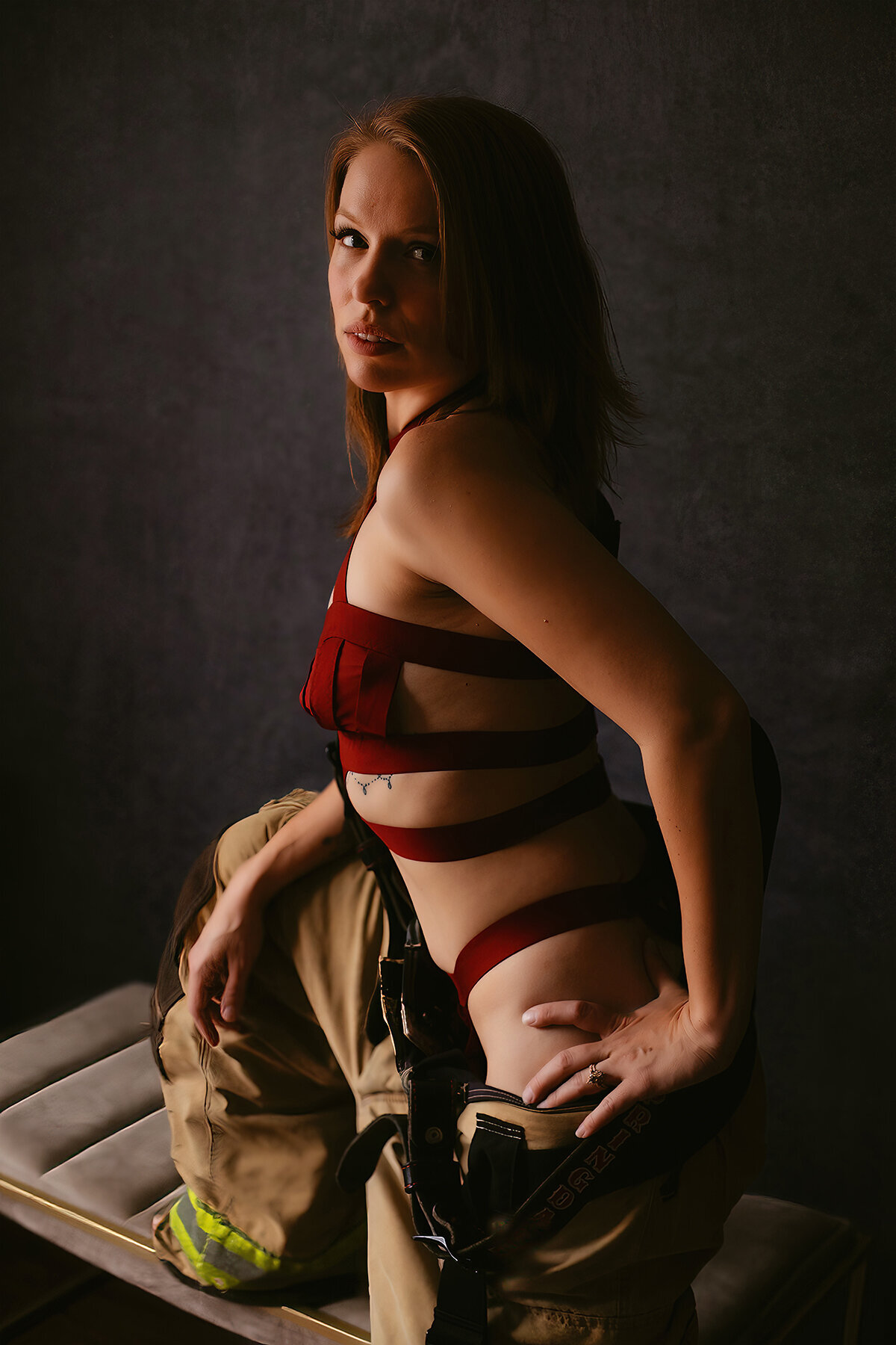 A woman in strappy red lingerie poses while wearing fireman gear during a Bentonville boudoir photoshoot.