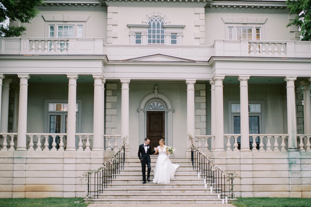 Bride in a white gown and groom in a black tux walking down the steps of the Great Marsh Estate while holding hands and smiling.