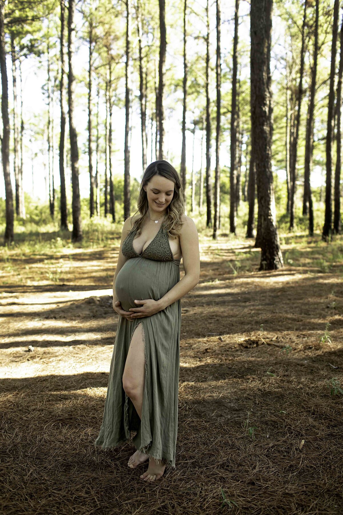 A serene maternity photo of the expectant mom standing alone in a tranquil pine tree forest, gently cradling her belly with a peaceful expression, captured by Chunky Monkey Photography.