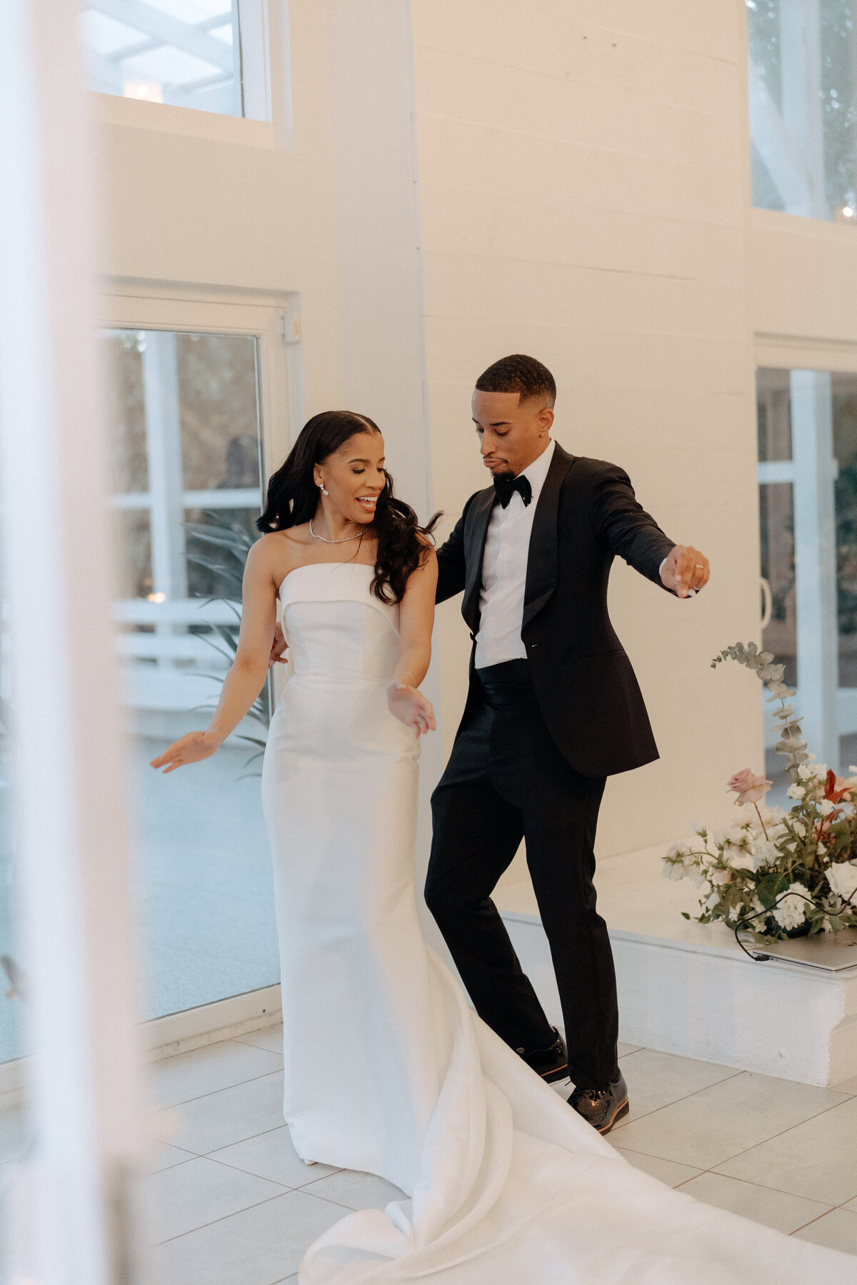 Bride and groom dancing at an indoor wedding reception in a modern white venue—strapless satin gown and black tux—Houston.