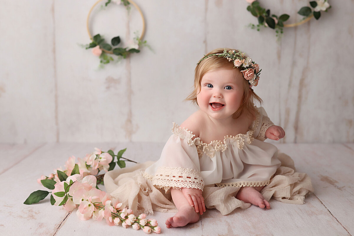 smily 1 year old sitting on a white wooden floor with flowers around