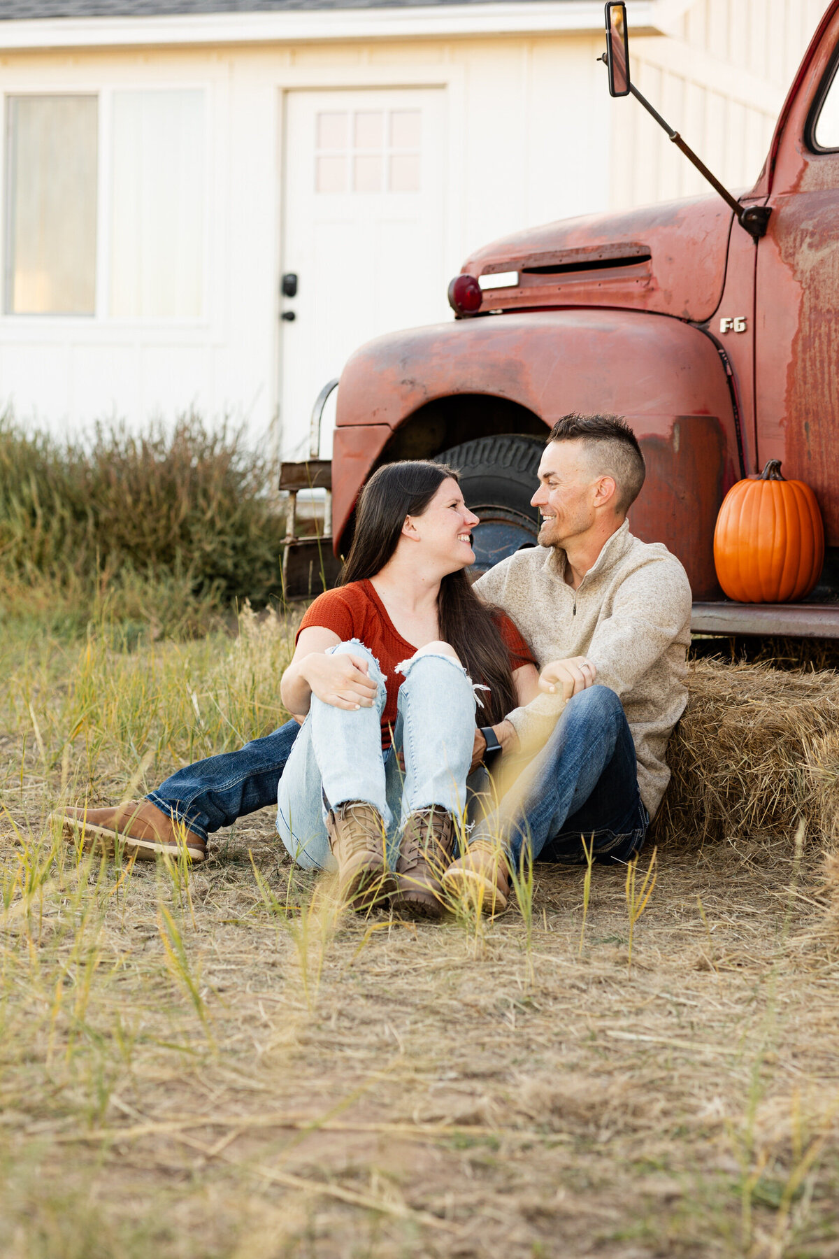 Husband and wife sit in the grass in front of a vintage red farm truck and smile at each other.