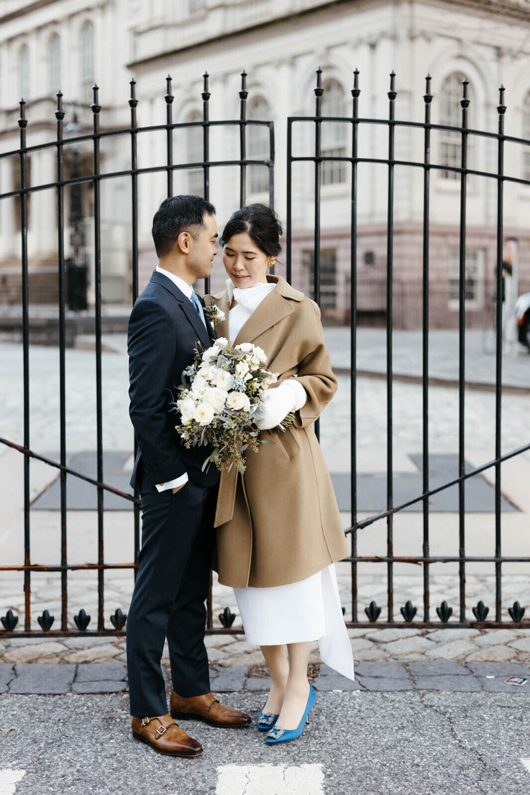 The bride, holding her flower bouquet, together with the groom, are standing in front of the gate of an old building.