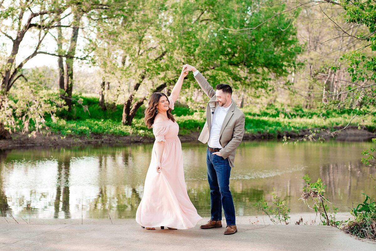 The groom, who is wearing a tan sports jacket, white shirt, dark wash jeans and a brown belt and shoes had one hand in his pocket and the other held over his head as he spins the bride. The bride is wearing a blush pink boho dress with short puffed off the shoulder sleeves and an empire waist. they are both smiling and laughing as they stand beside a pond with lush greenery and trees.