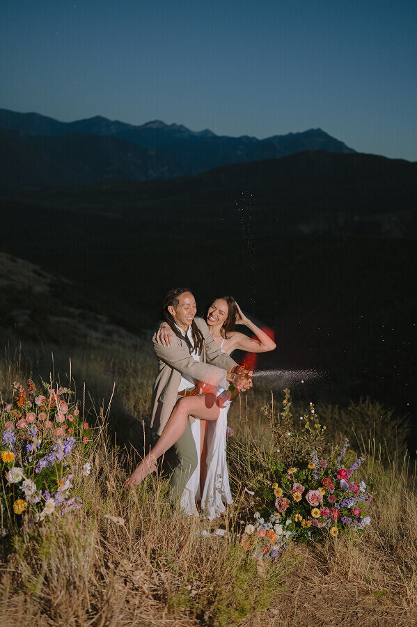 A flash photo of a couple on their elopement day at night. A woman is popping a bottle of champagne as her wife holds onto her.