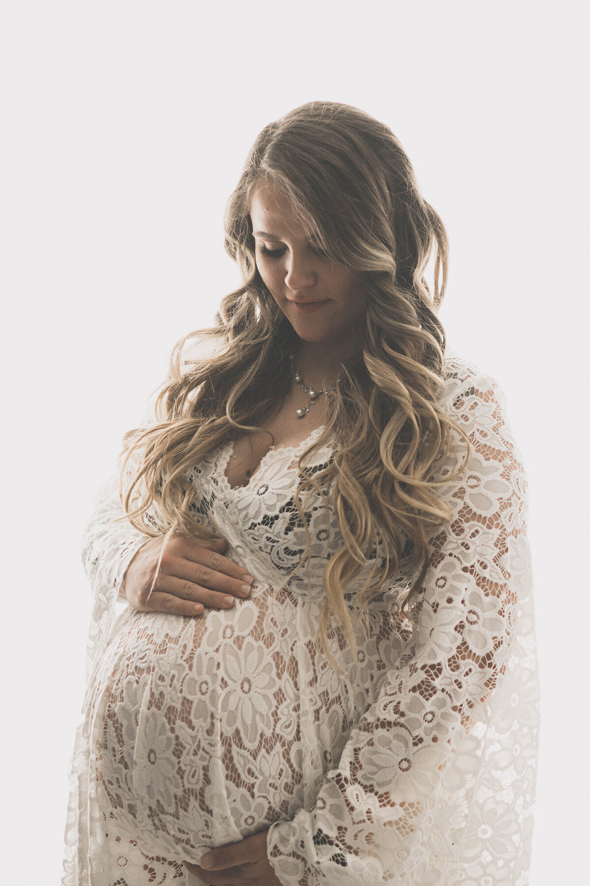 A mom to be stands in a studio smiling down at her bump in her hands in a white lace maternity gown