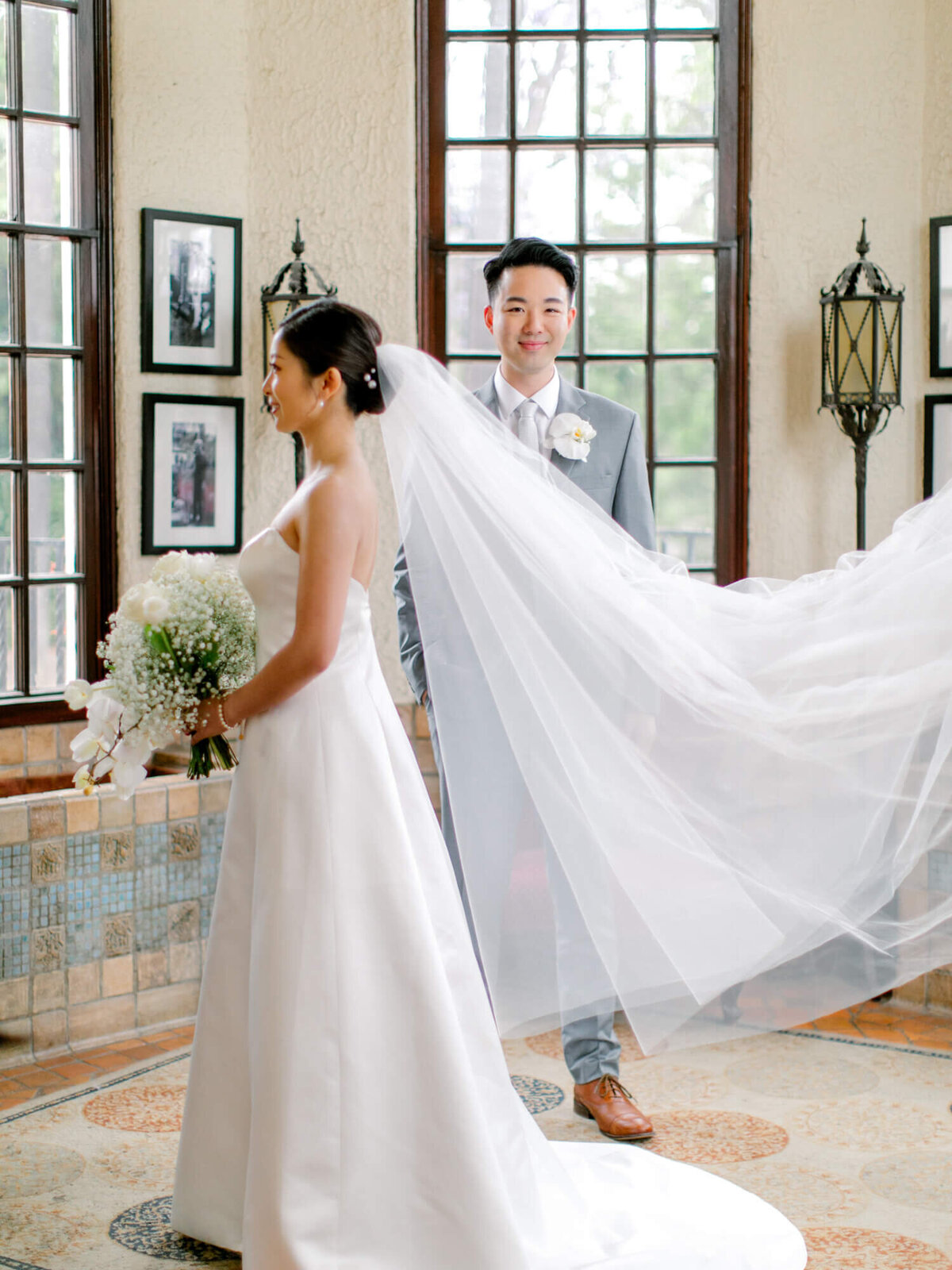 A bride in a white dress stands smiling with a floral bouquet, while her veil flows behind. A groom in a gray suit stands behind her, smiling. The room has large windows and framed photos, creating a romantic atmosphere.
