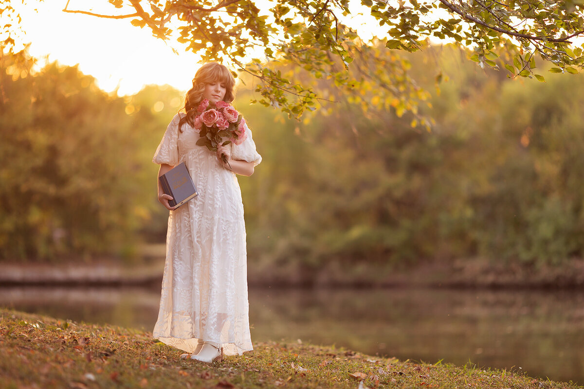 Dallas senior photographer captures a young woman in a white dress holding a bouquet of pink roses and a book during sunset by the water.