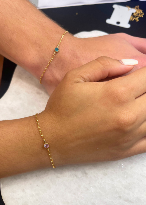 A close-up of two hands wearing matching gold permanent bracelets with birthstone charms, symbolizing a meaningful connection.