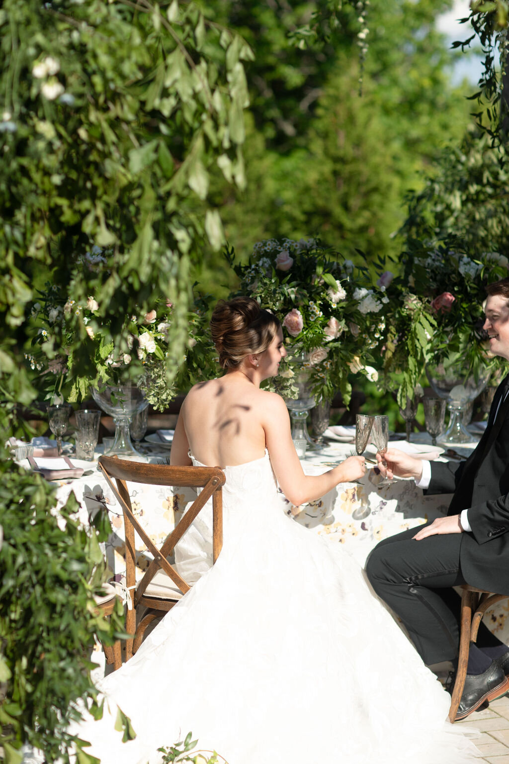 a bride in a white gown and groom in a black suite sit under a pergola or beautiful greenery on crossback chairs at Willowbrook wedding venue