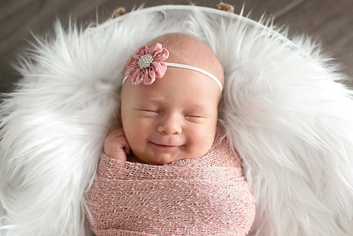 Newborn girl smiling while sleeping wrapped in a pink swaddle with matching headband.