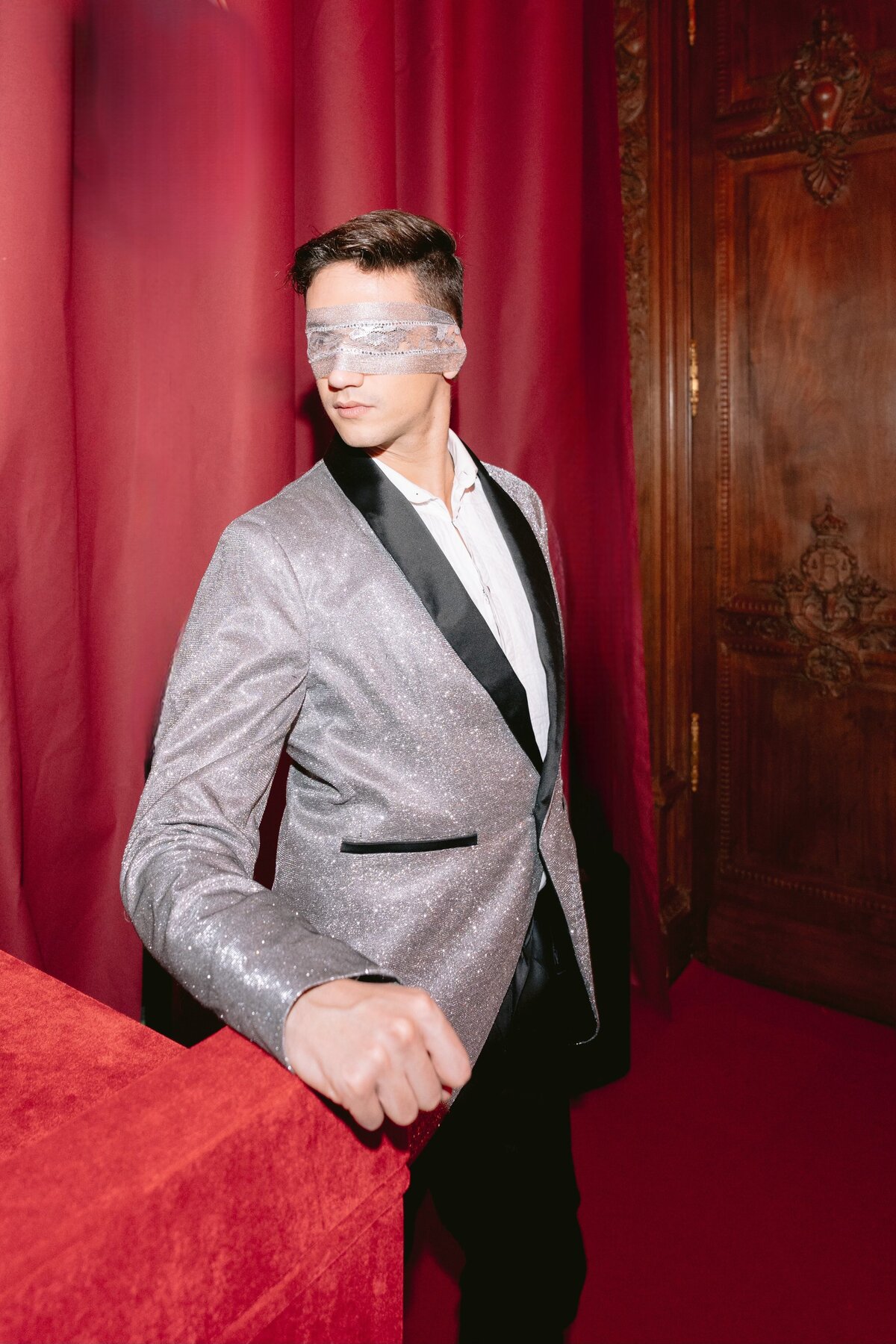 The photo was taken at the prestigious Shangri La Hotel & Ritz Paris Hotel in Paris, France, capturing the elegant ambiance of the location. Talented photographer Matteo Coltro expertly framed the shot, highlighting the luxurious setting. Additionally, Zen Film Works beautifully captured the essence of the moment through videography.  In the image, a man in a shimmering silver jacket stands confidently, his right arm casually resting on a lavish red velvet surface. He is adorned in a crisp white shirt, adding a contrast to the metallic hue of his outfit. Notably, his eyes are covered by a striking blindfold made of intricate silver mesh, adding an element of mystery to the composition.  The backdrop of the photograph is adorned with opulent rich red curtains and a wooden door displaying elaborate carvings, enhancing the overall sophistication of the scene. Each detail in the image contributes to a sense of refinement and luxury that is synonymous with the prestigious hotels where the photo was taken.