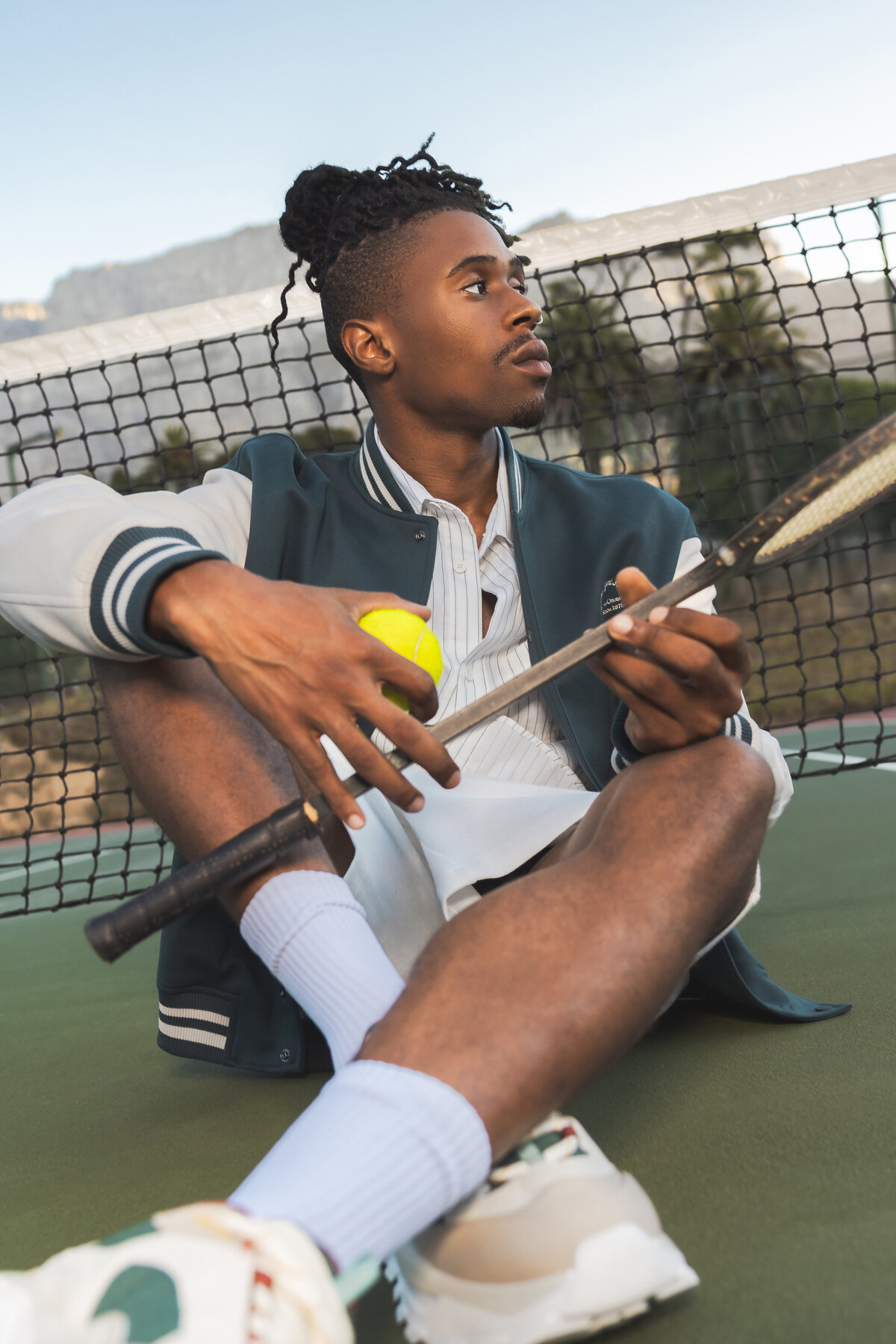 Man sitting on a tennis court in front of the net, holding a tennis ball and racket, wearing a varsity jacket and looking to the side with Table Mountain in the background.