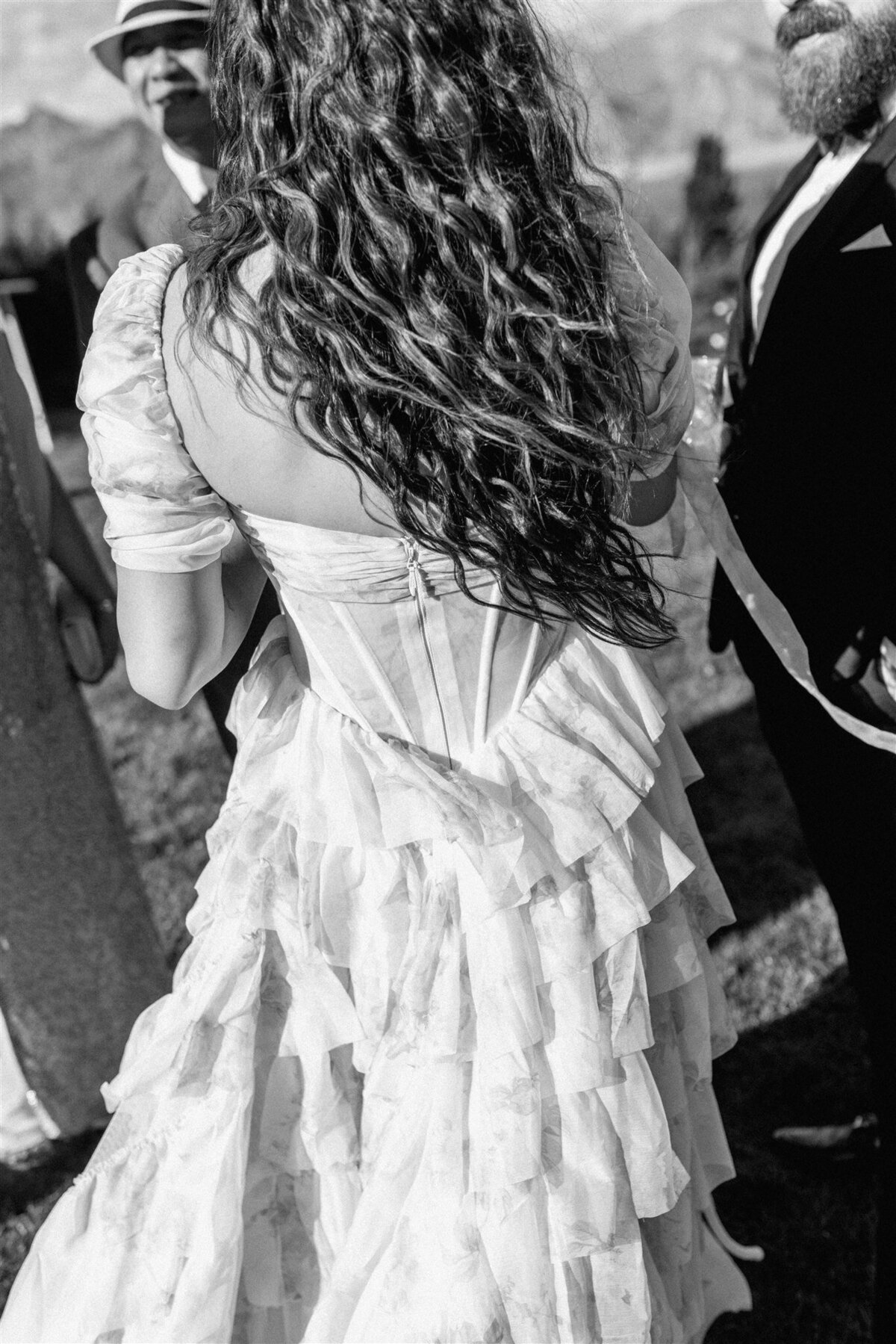 Black and white photo of bride’s back showcasing cascading curls and ruffled gown details during outdoor cocktail hour in Lake Hāwea.