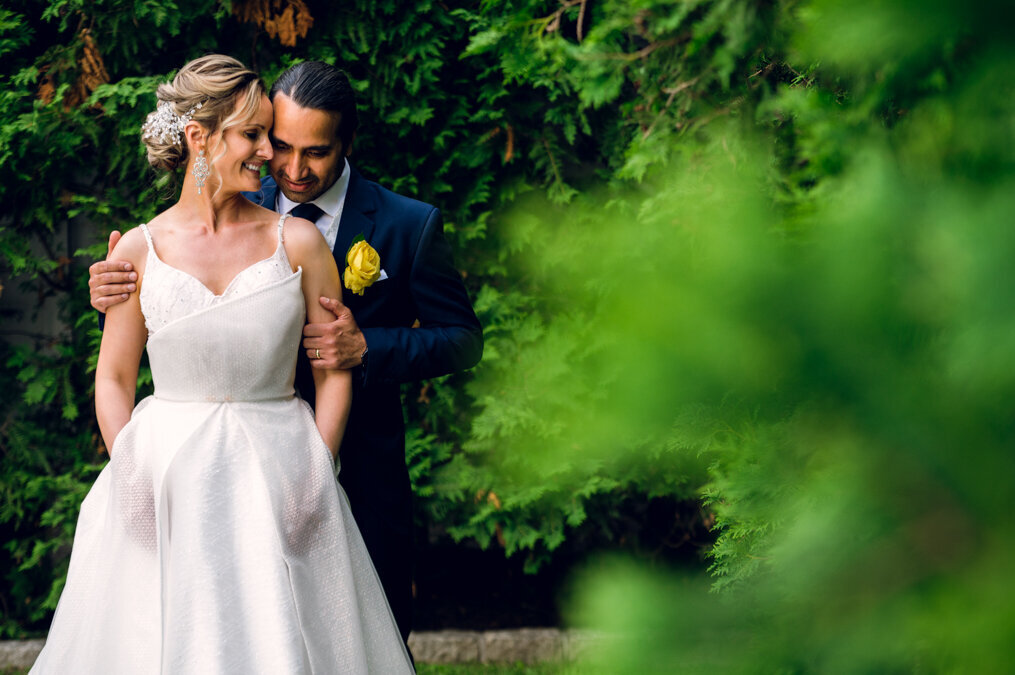 A bride and groom embracing in front of greenery.
