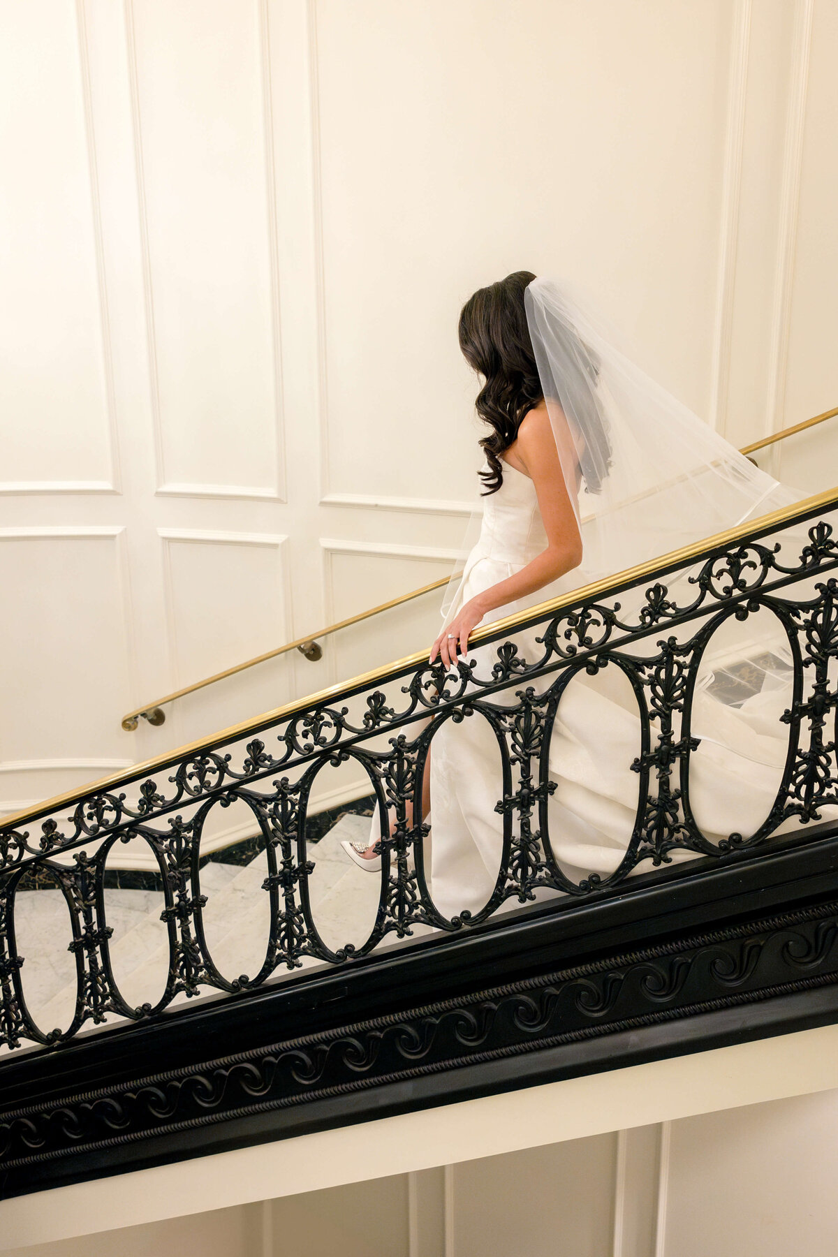 A bride in a white wedding dress and veil descends a staircase with ornate wrought iron railing, her face turned away. The setting is elegant with cream-colored walls and a gold handrail.