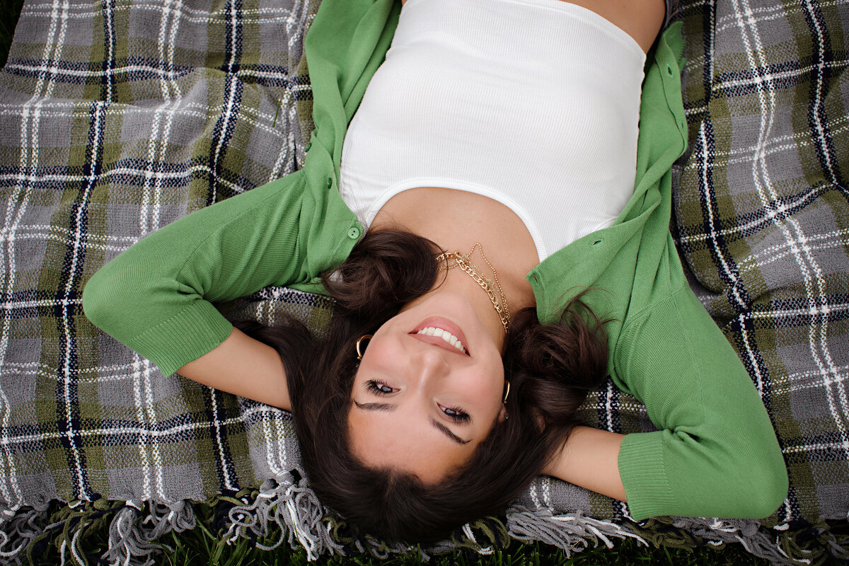 Editorial-style portrait of high school senior in green cardigan and white tank top, laying on a blanket with greenery all around.