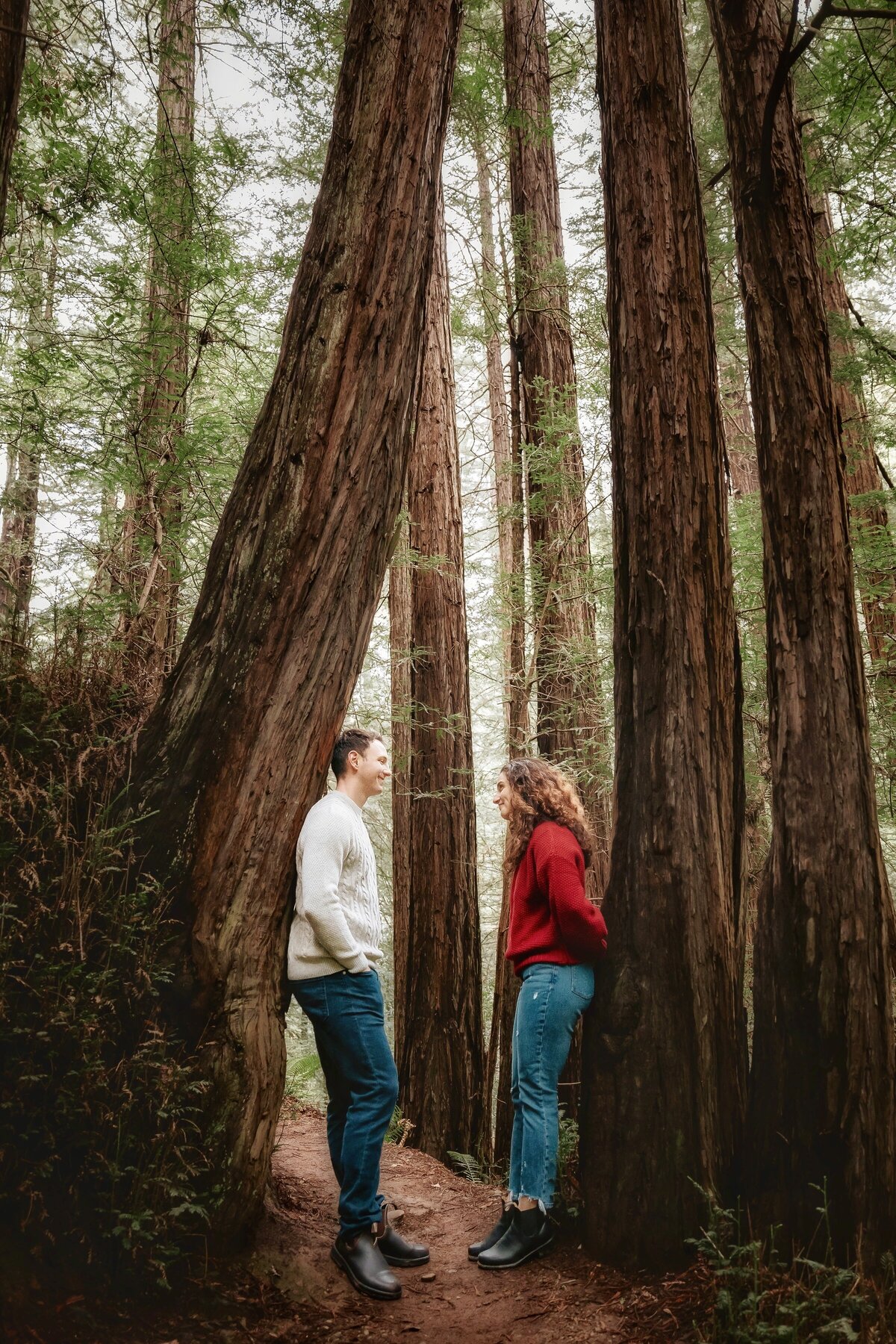 Couple look at each other while leaning on redwood trees in Muir Woods