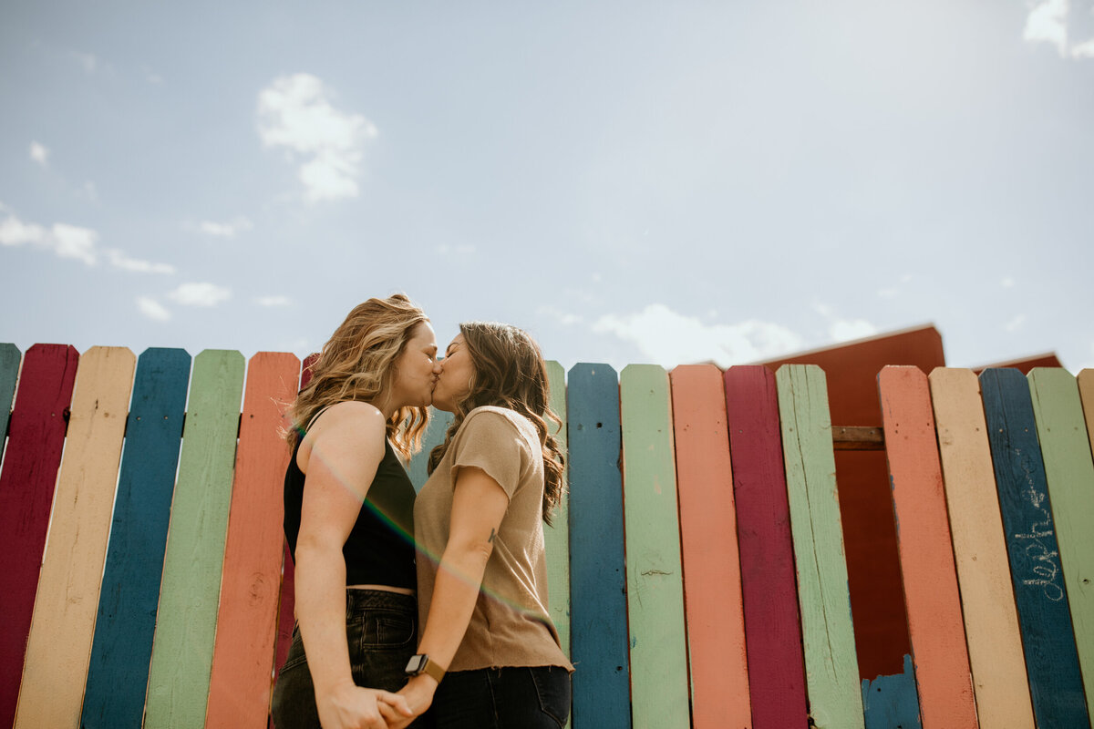 same sex couple kissing in front of a rainbow fence