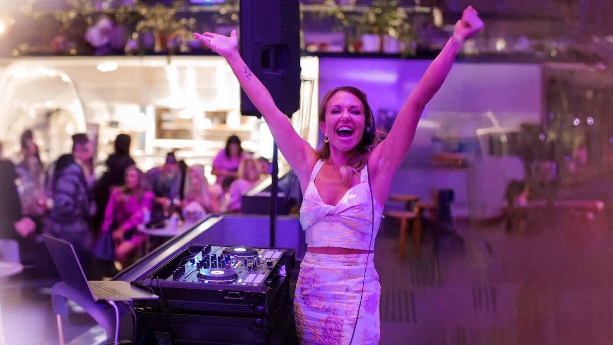 Smiling female DJ with arms raised in celebration behind a DJ booth, performing at a vibrant indoor event with a lively crowd and purple lighting.