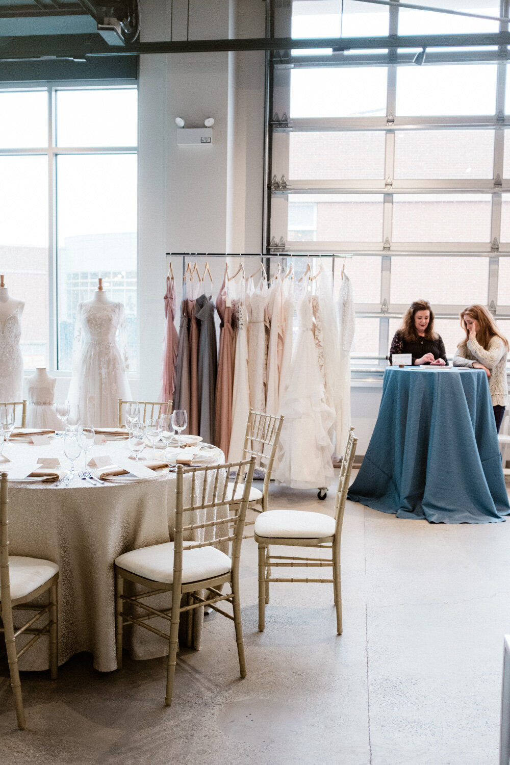 Two girls standing beside round table with blue cloth, wedding gowns hanging, and wedding tables and chairs