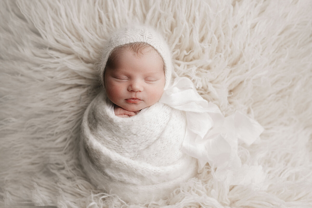 Studio newborn photography - Newborn baby girl wrapped in a white knit swaddle on a white flokati. Baby is wearing a knit bonnet with long ribbon ties flowing to the right of the frame.