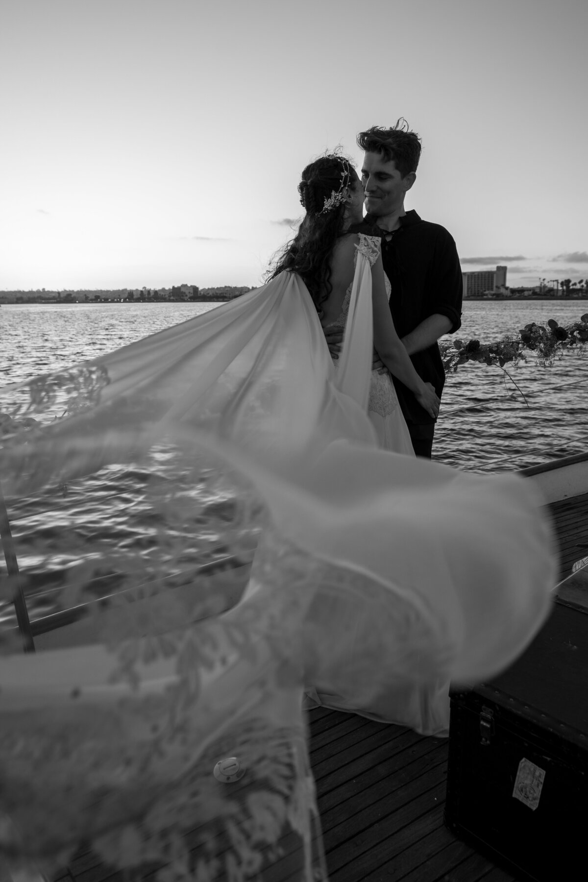 Bride and groom on yacht in San Diego in black and white embracing each other while her Claire Pettibone chiffon gown is blowing in the ocean breeze