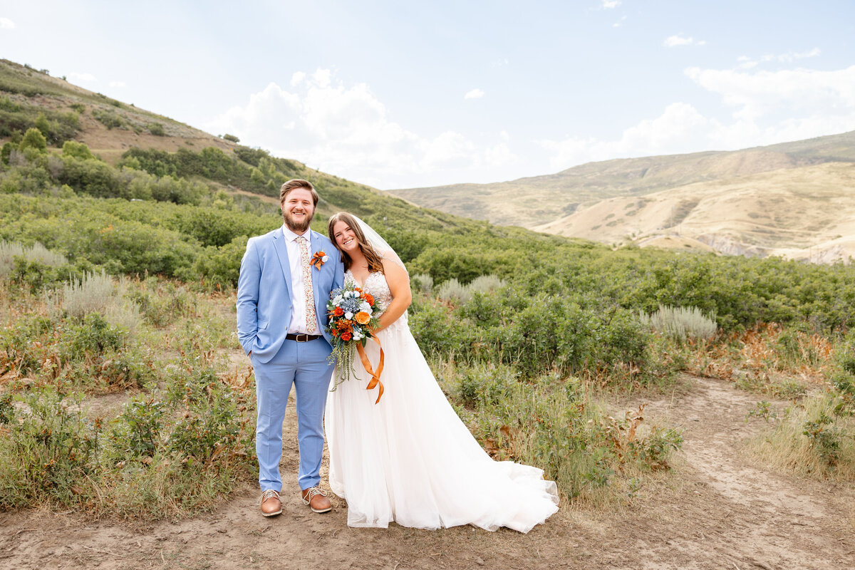 A bride in a flowing white gown and a groom in a light blue suit stand together in a scenic mountainous landscape in Provo Canyon, Utah. The bride holds a bouquet with blue and orange flowers, and both smile at the camera.