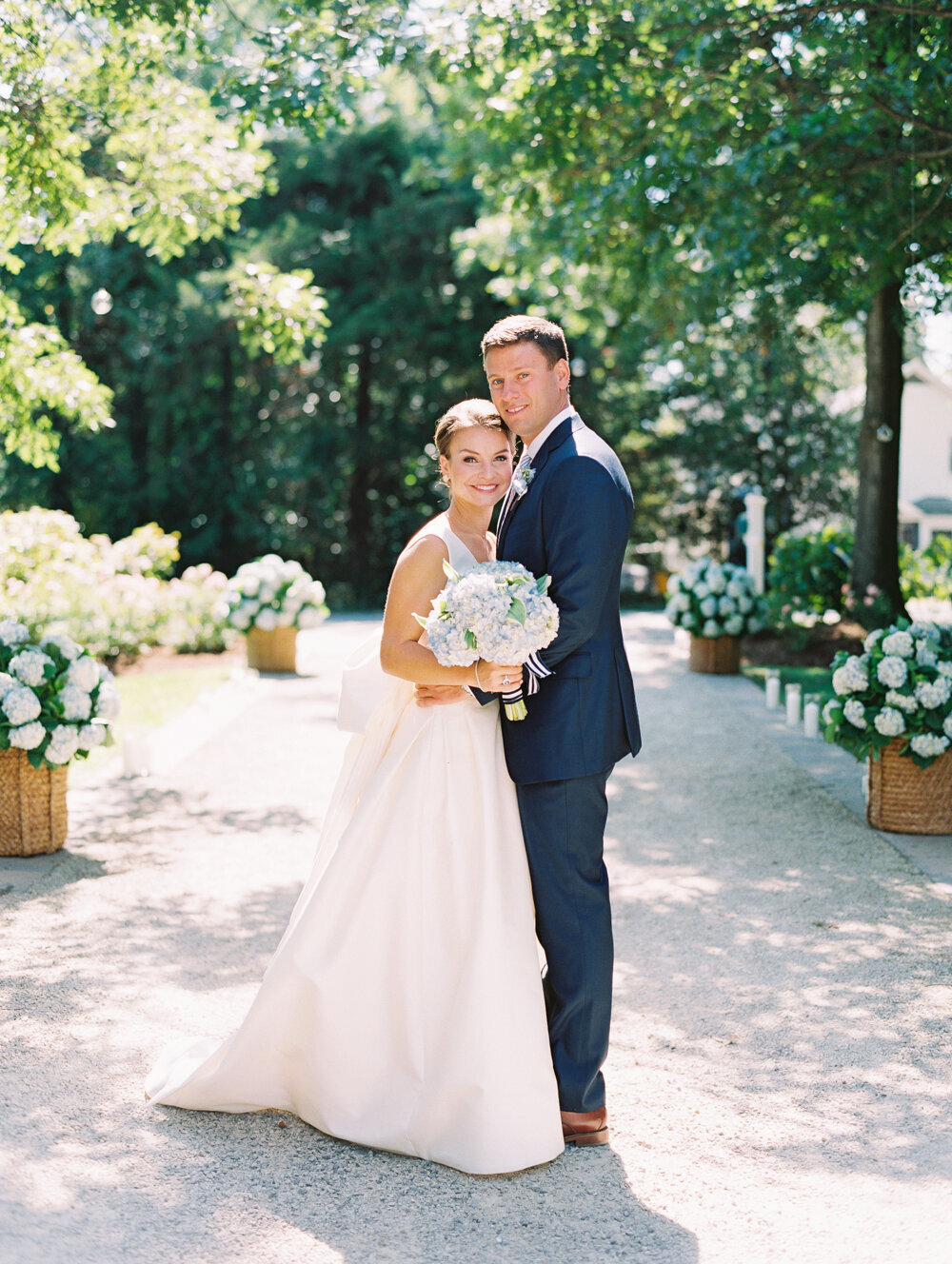 A smiling bride in a white gown and groom in a navy blue suit stand close together outdoors, holding a bouquet of light blue flowers. They are surrounded by lush greenery and white floral arrangements, with sunlight filtering through the trees.