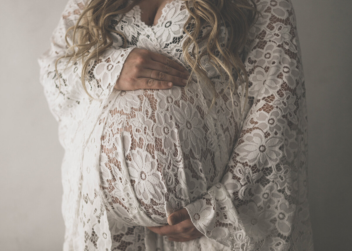 Details of a mom's hands on her bump in a white lace maternity gown