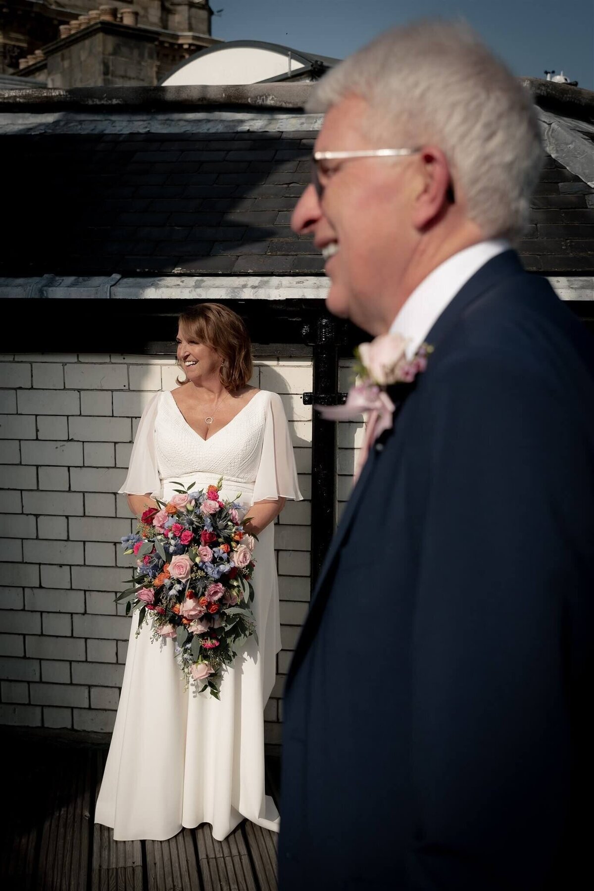 A couple stand apart on the roof of the Balmoral Hotel in Edinburgh. The bride in background holding a bouquet of flowers, the groom slightly out of focus in the foreground.