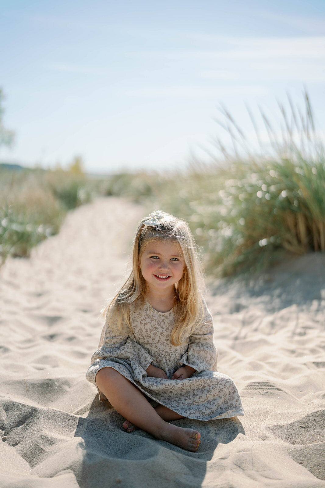 A young girl smiling while sitting on the sandy path surrounded by dune grass at Weko Beach in Bridgman Michigan.