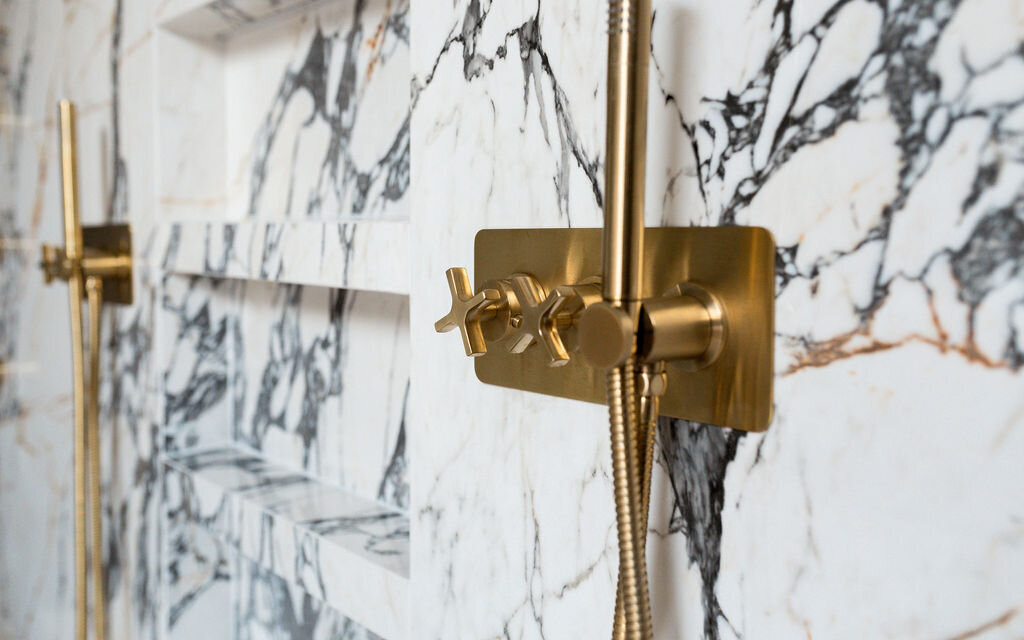 Two modern gold shower fixtures mounted on a white marble wall with black veining. Between them, multiple shelves are built into the marble wa