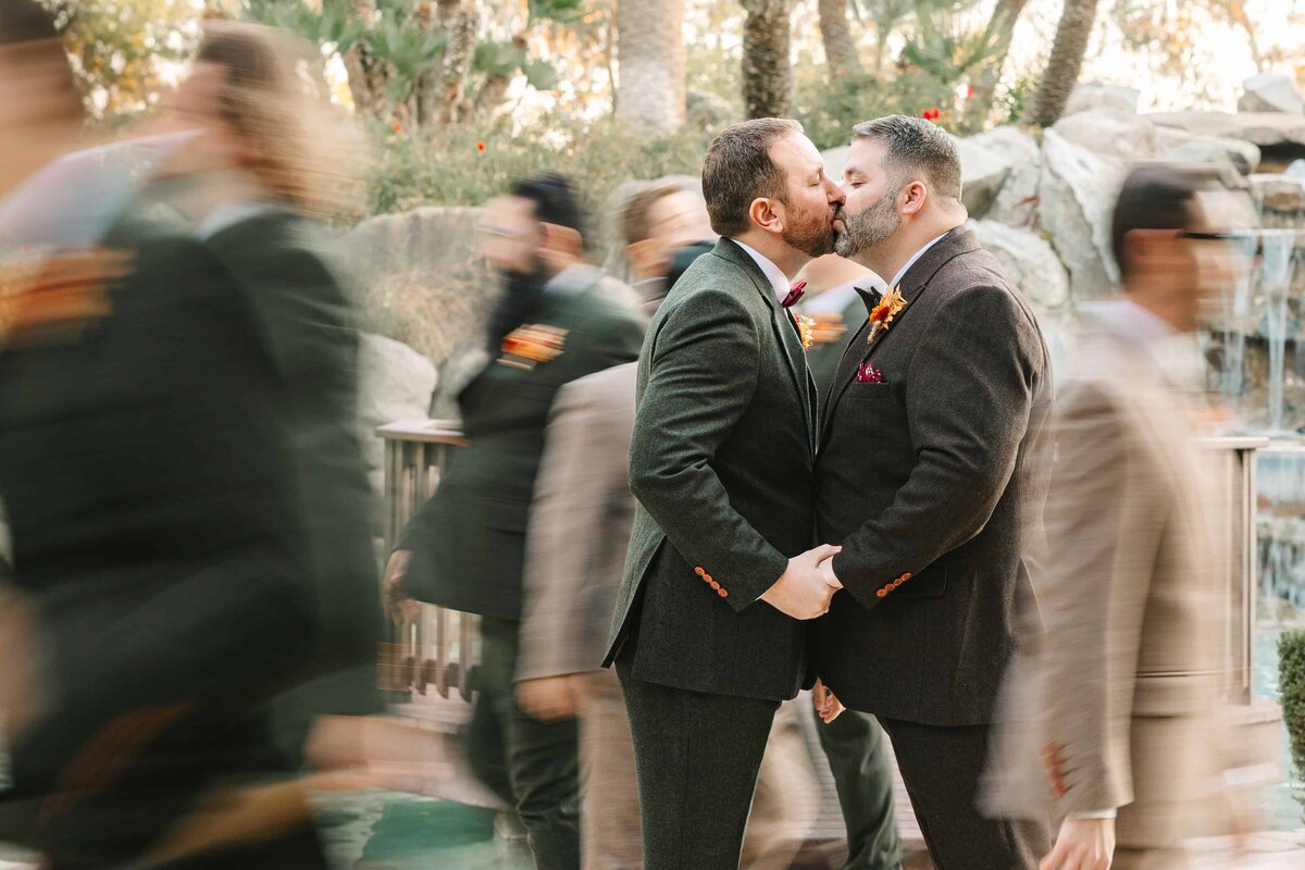 two grooms kissing as their wedding party rushes by in a blur in the background