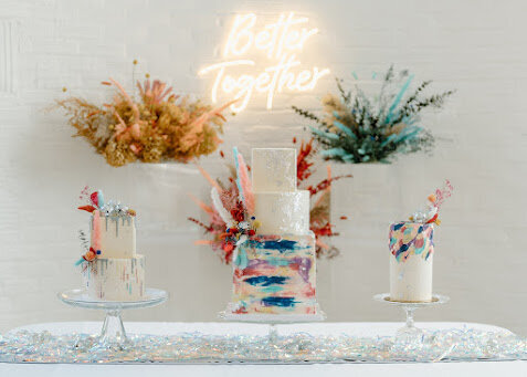 Photo shows three cakes on a table with a silver sequined table runner. The middle cake is three tiers with the bottom tier having different shades of water color style frosting, and the top two being white with silver leaf. The cake on the left is a two tier cake with a silver drip and colorful dot stenciling. The right cake is a tall single tier cake with mostly white frosting, and swatches of the same colors on the top in a random pattern. All have mini disco balls and colorful dried floral accents.