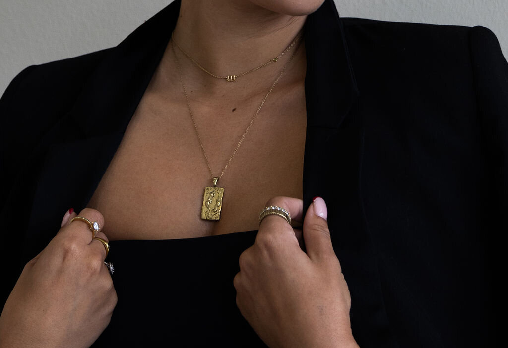 Close-up of a woman wearing a black suit with the jacket draped over her shoulders, complemented by a gold necklace and rings, exuding elegance and sophistication.