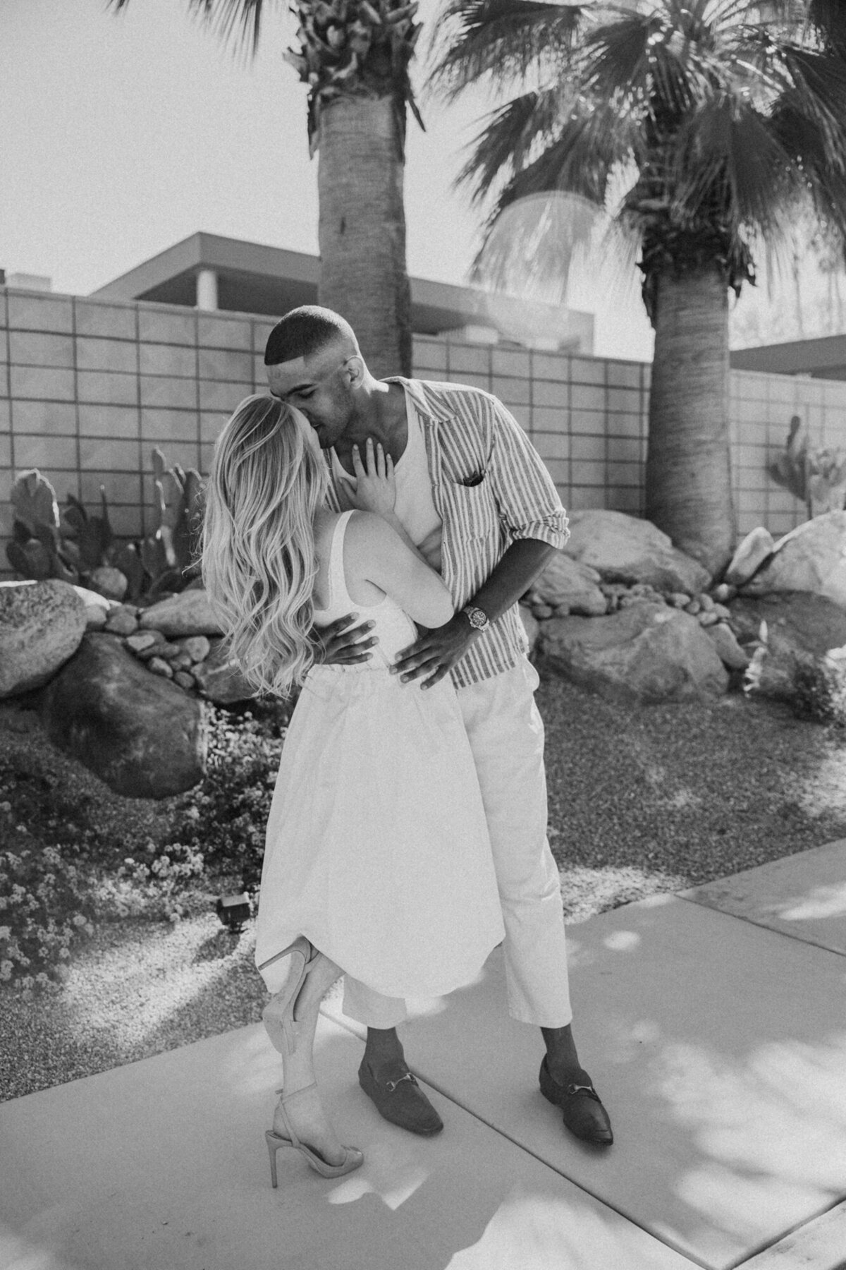 Black and white candid image of couple smiling together, holding hands in a garden pathway.