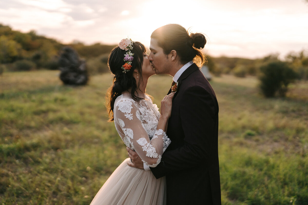 A photograph in color of Arista and Sean on their wedding day at Old Glory Ranch in Wimberley, near Austin, Texas. The couple are standing in a field at sunset with the sun glowing from behind them. They are facing each other and kissing. The bride has a floral crown in her dark hair and she is wearing a long sleeved lace blouse with a flowing skirt. Wedding photography by Stacie McChesney/Vitae Weddings.