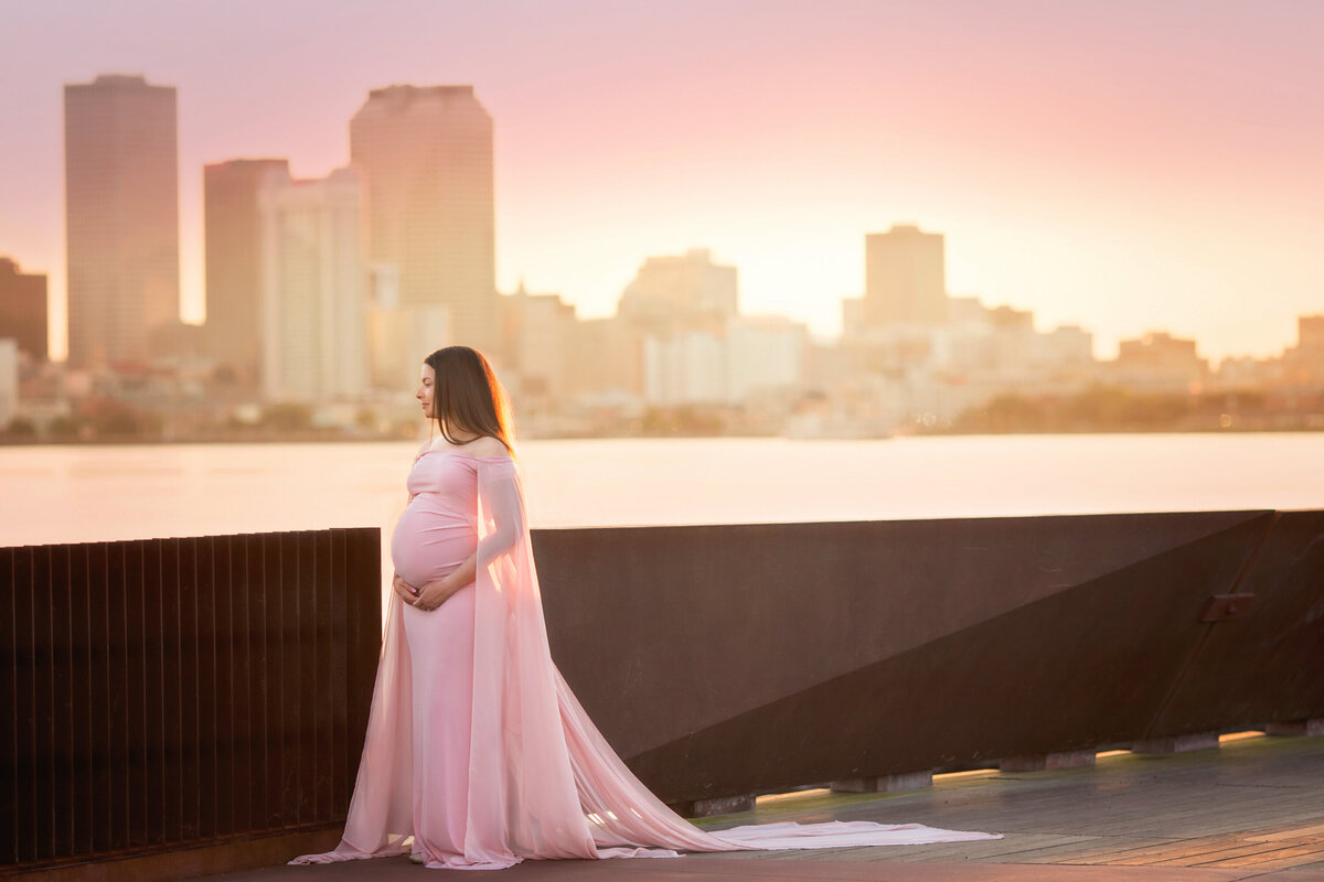 Black woman in blush Silk Fairies Maternity dress holding her pregnant belly.  The dress has long, chiffon sleeves.  She is standing in Crescent Park and has the New Orleans skyline behind her.  There is a pink sunset.