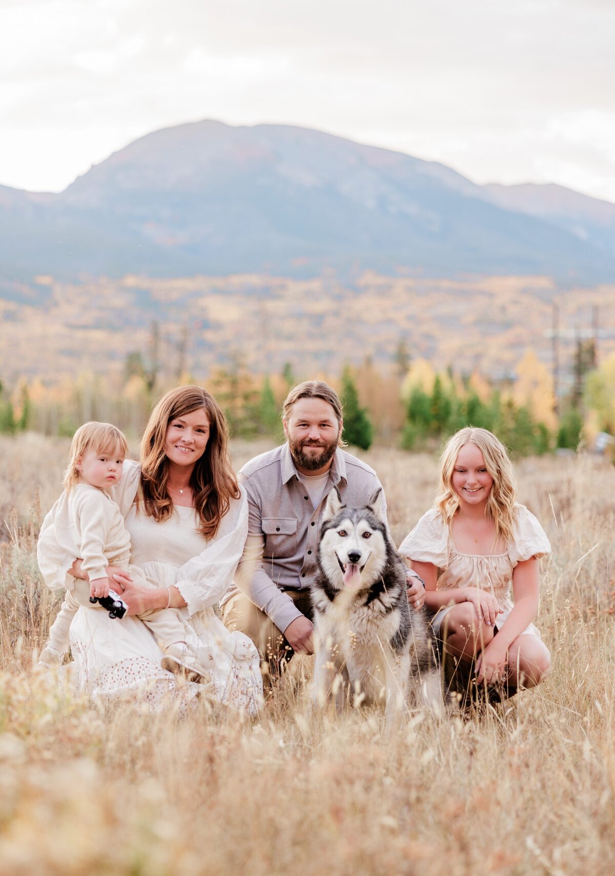 A family of 4 smile while kneeling in tall golden grass behind sitting husky