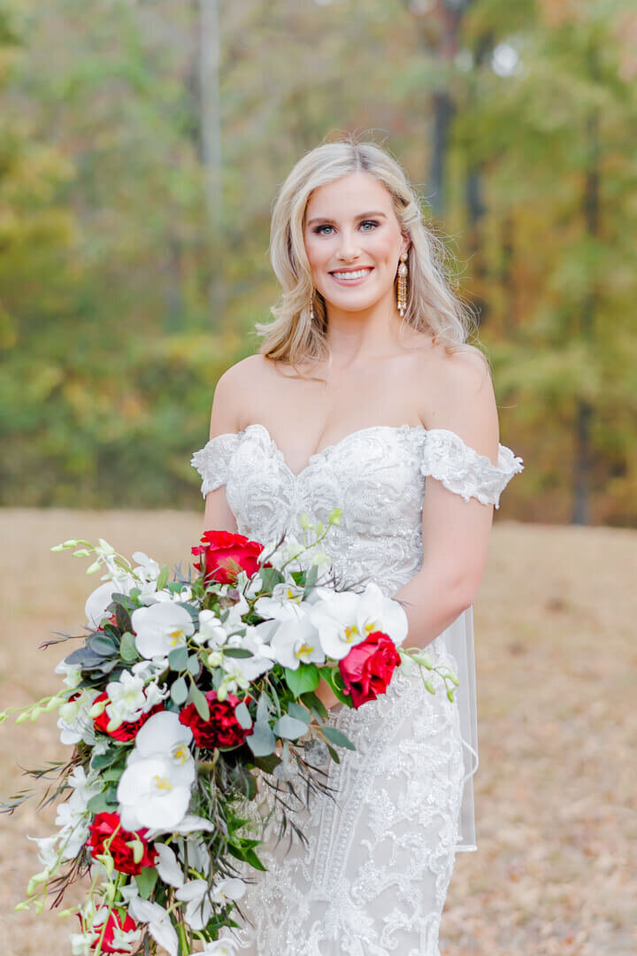 An image taken by an Arkansas wedding photographer of a bride standing by a forest holding a large red and white bouquet