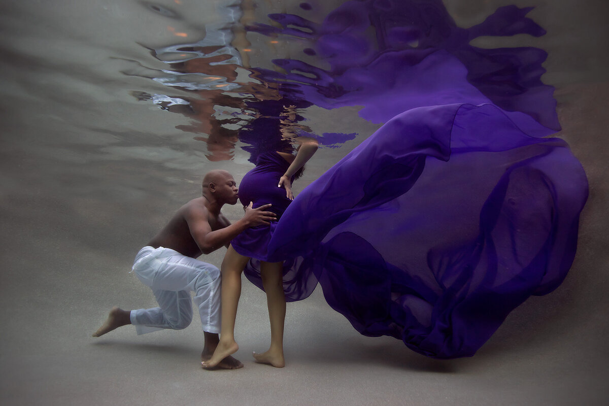 A heartfelt underwater maternity photo showing a loving father gently holding his partner's belly, surrounded by the flowing purple fabric of her gown. This stunning underwater maternity photoshoot beautifully captures their bond.