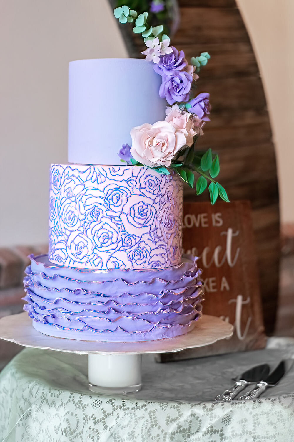 Lavender and pink 3 tier wedding cake. Bottom tier has lavender ruffles edged in pink; middle tier is pink with lavender rose stencil; top tier is lavender with cascading pink and lavender roses,  ruscus and eucalyptus leaves