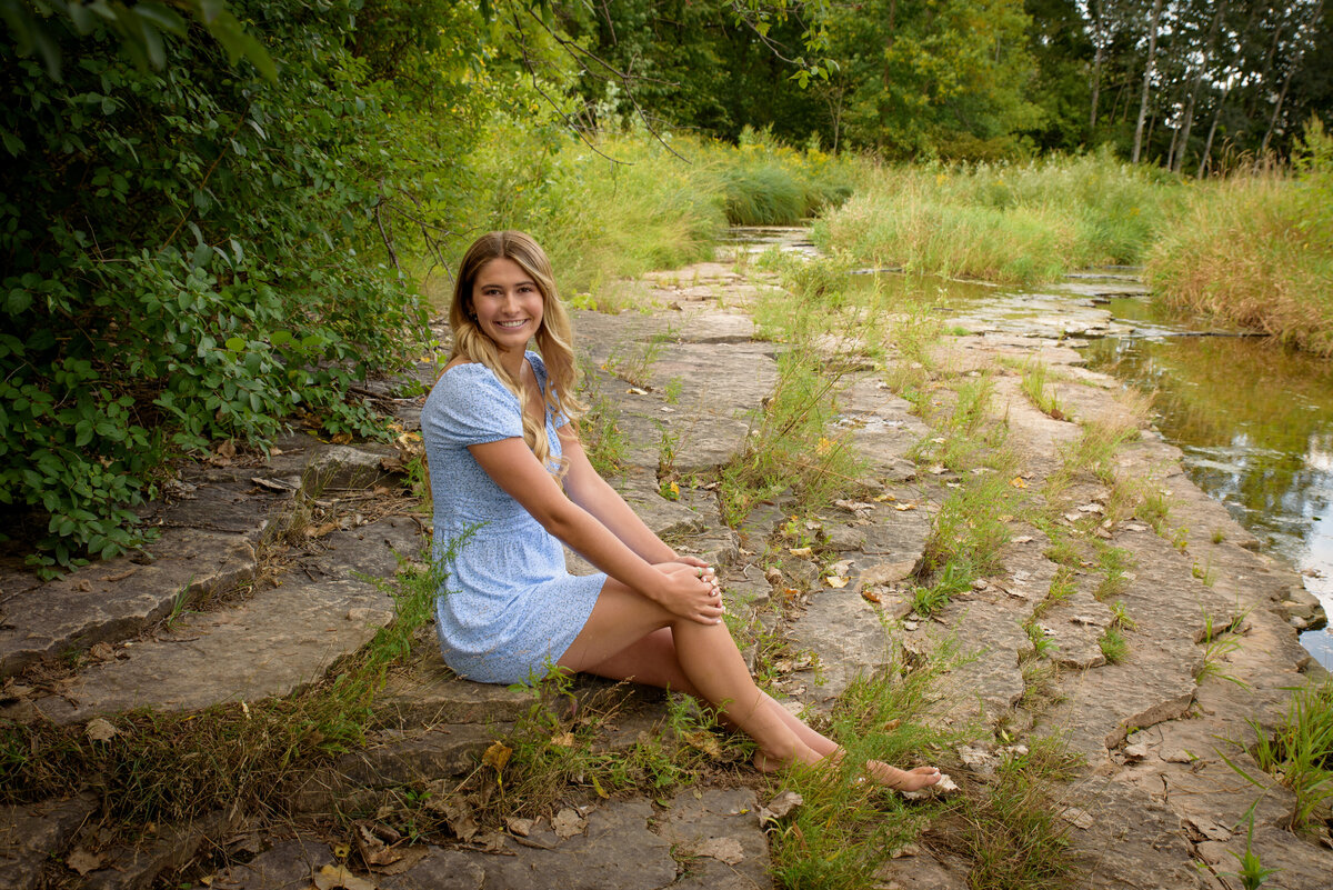 De Pere High School senior girl sitting on edge of creek wearing a light blue summer dress at Fonferek Glen County Park in Green Bay, Wisconsin