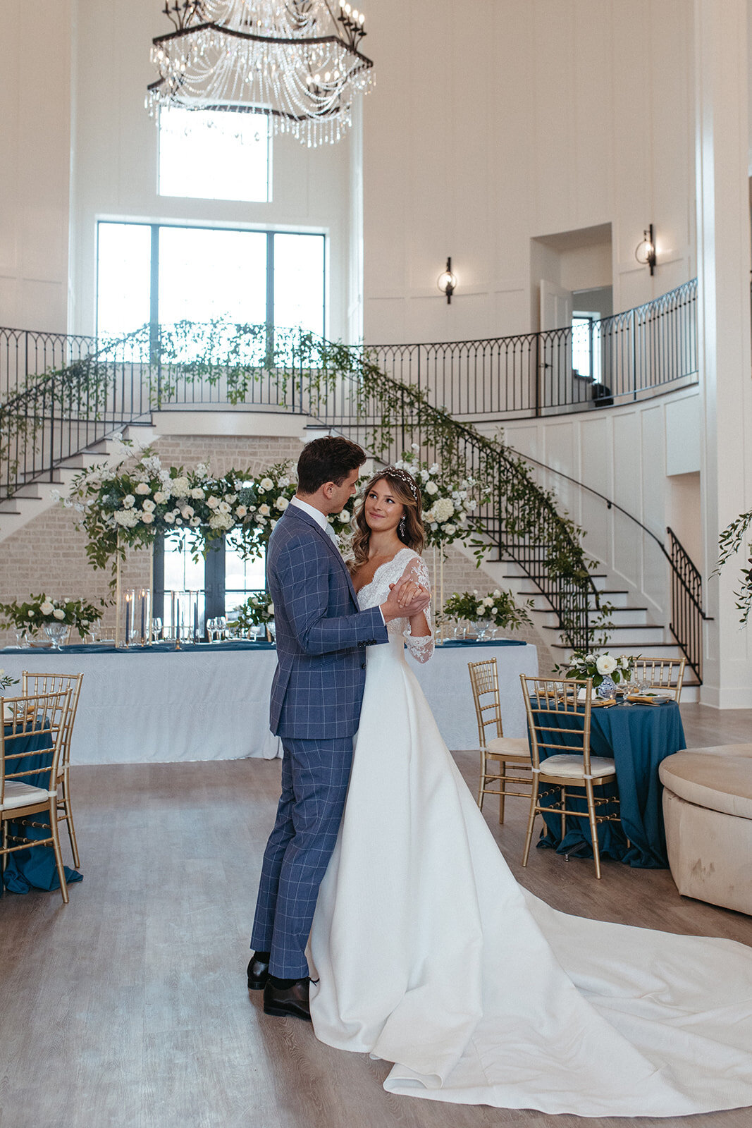 Bride and groom in a blue suit and white wedding gown dancing in a banquet room with white flowers and greenery.