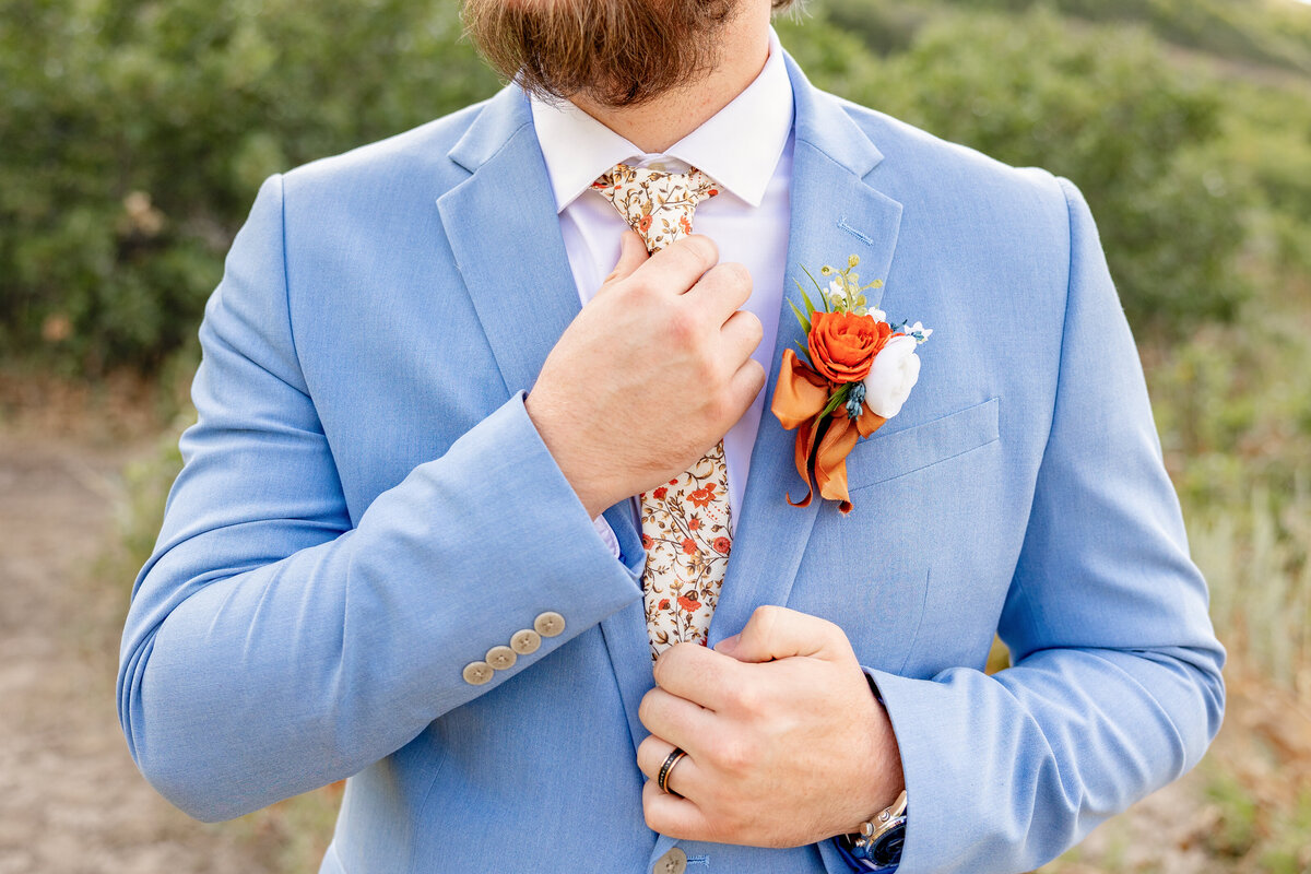 A close-up shot of the groom adjusting his floral-patterned tie, highlighting his light blue suit and an orange and white boutonniere pinned to his lapel.