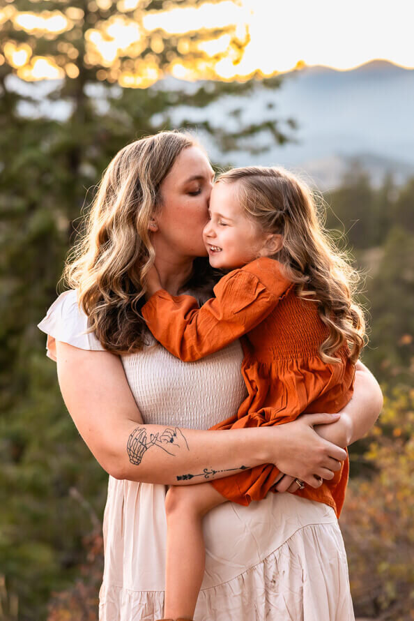 A mom and her small daughter hug with evergreen trees behind them.