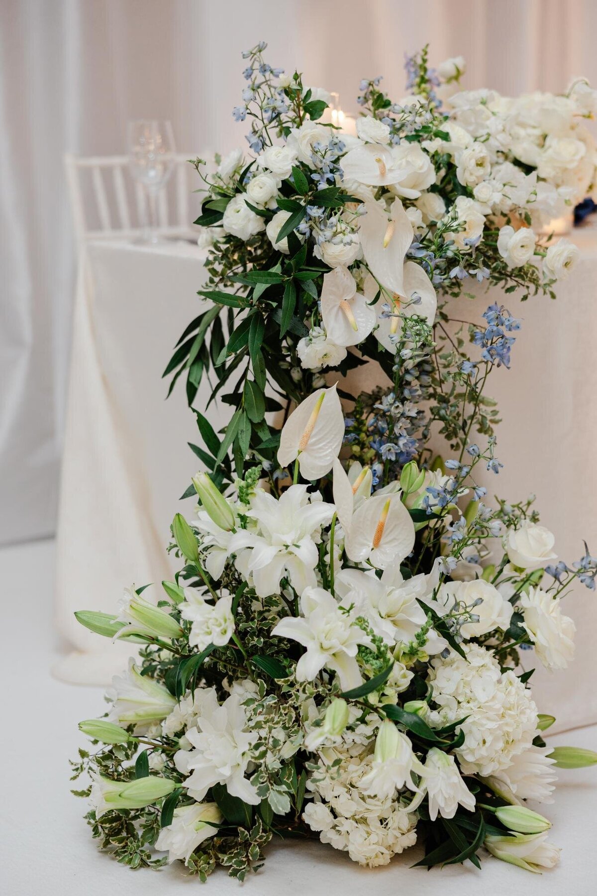 Elegant floral arrangement with white lilies, hydrangeas, and other greenery on a draped table at an event.