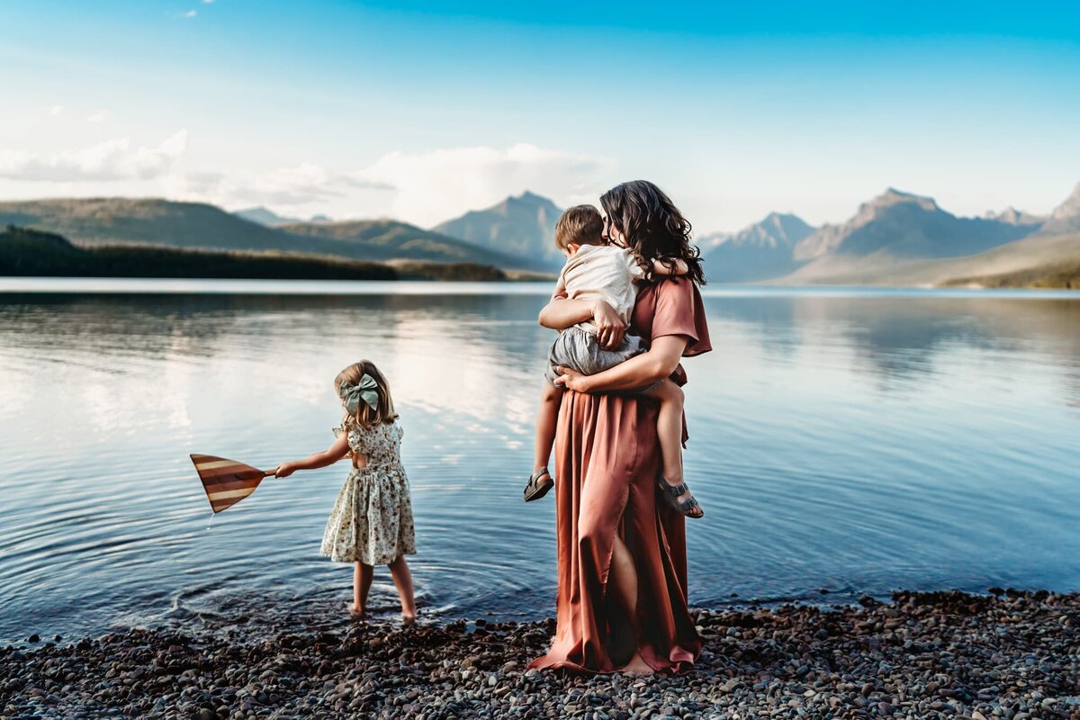 A mother holds her son on the shore of Glacier National Park's Lake McDonald while her daughter plays in the water nearby, during a vacation family photography shoot by Love Michelle Photography