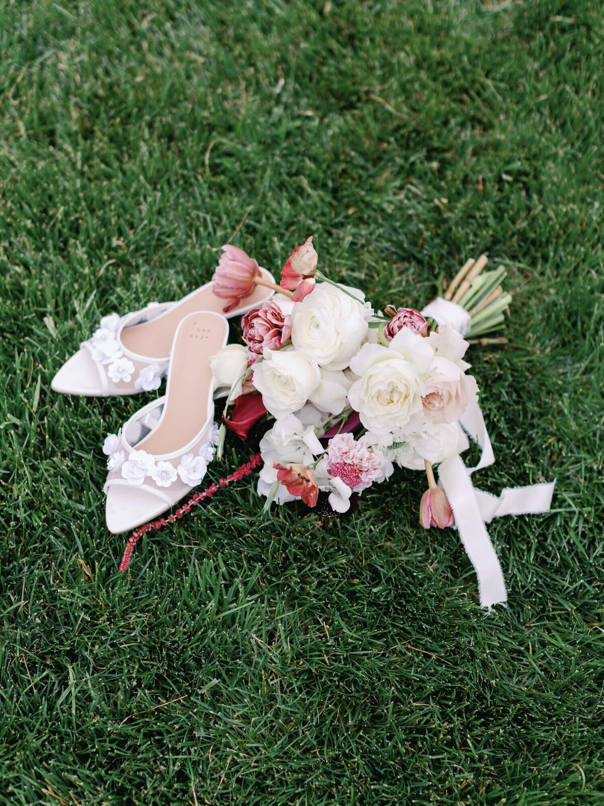 White bridal heels adorned with floral appliques lie on lush green grass beside a bouquet of pink and white roses and tulips.